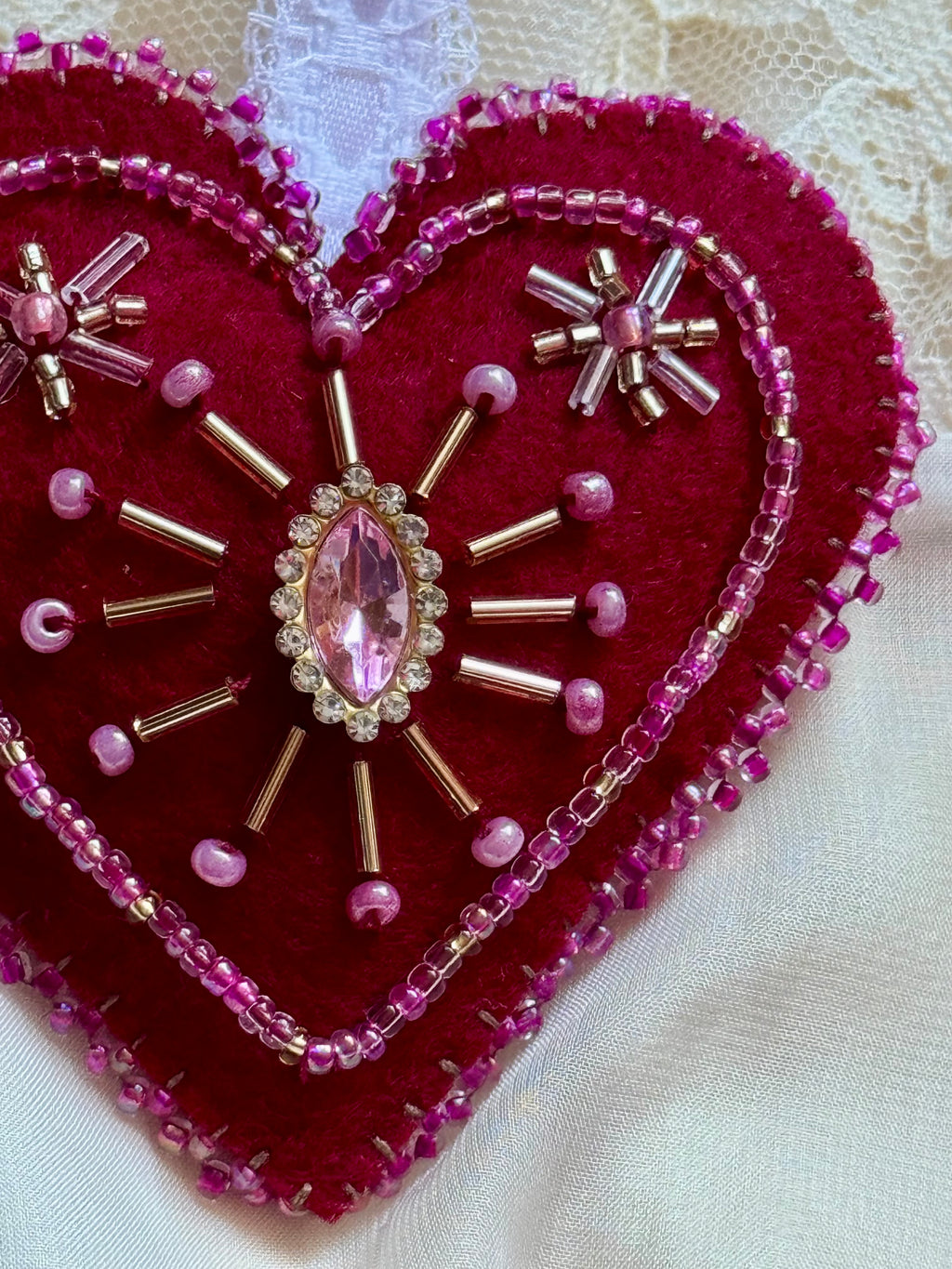Heart-shaped embroidered design with pink gemstone and beads on a white background