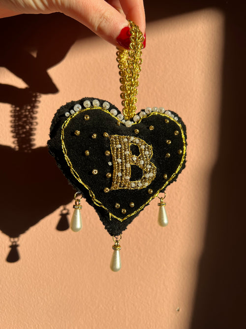 Decorative black heart ornament with gold accents, inspired by Anne Boleyn's 'B' necklace, held by a hand on a beige background
