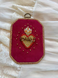 Embroidered wall hanging with a sacred heart design on velvet, framed in a wood-look frame with metal hanging, on a white fabric surface.
