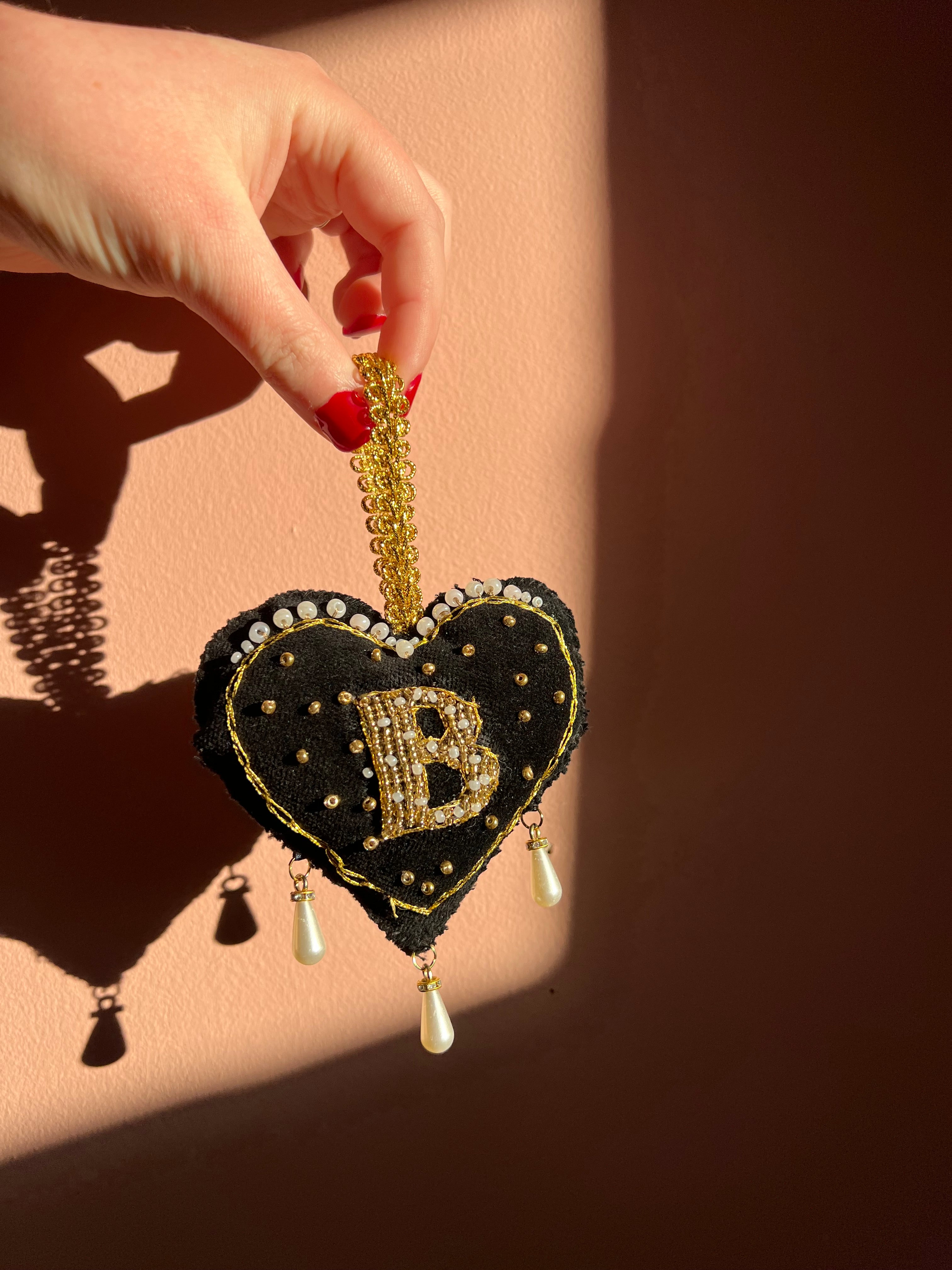 Decorative black heart ornament with gold accents and pearl details, inspired by Anne Boleyn's 'B' necklace, held by a hand on a beige background