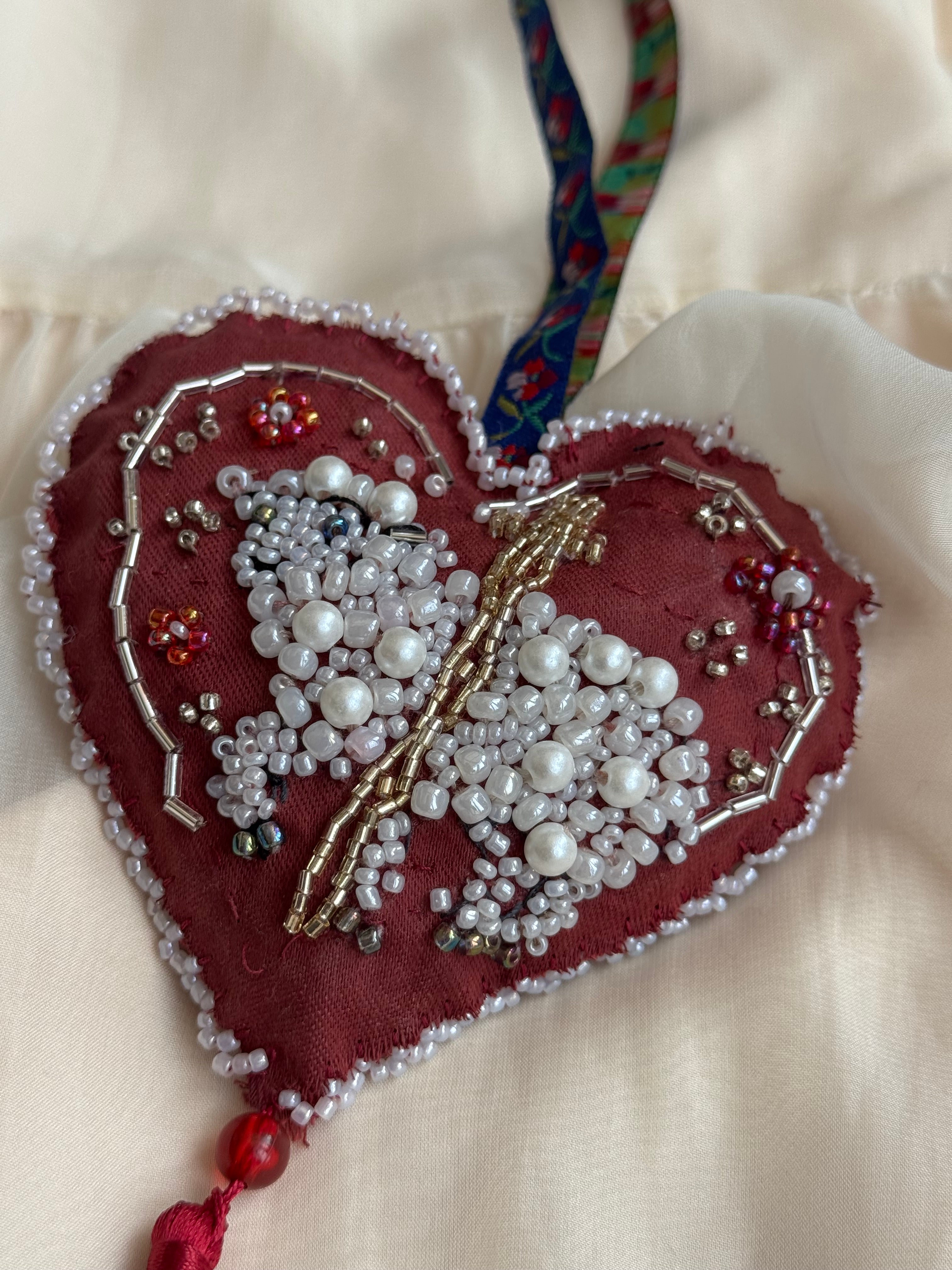 Heart-shaped ornament with beadwork shaping a sacred lamb on a white background with cross, floral hanger and tassle