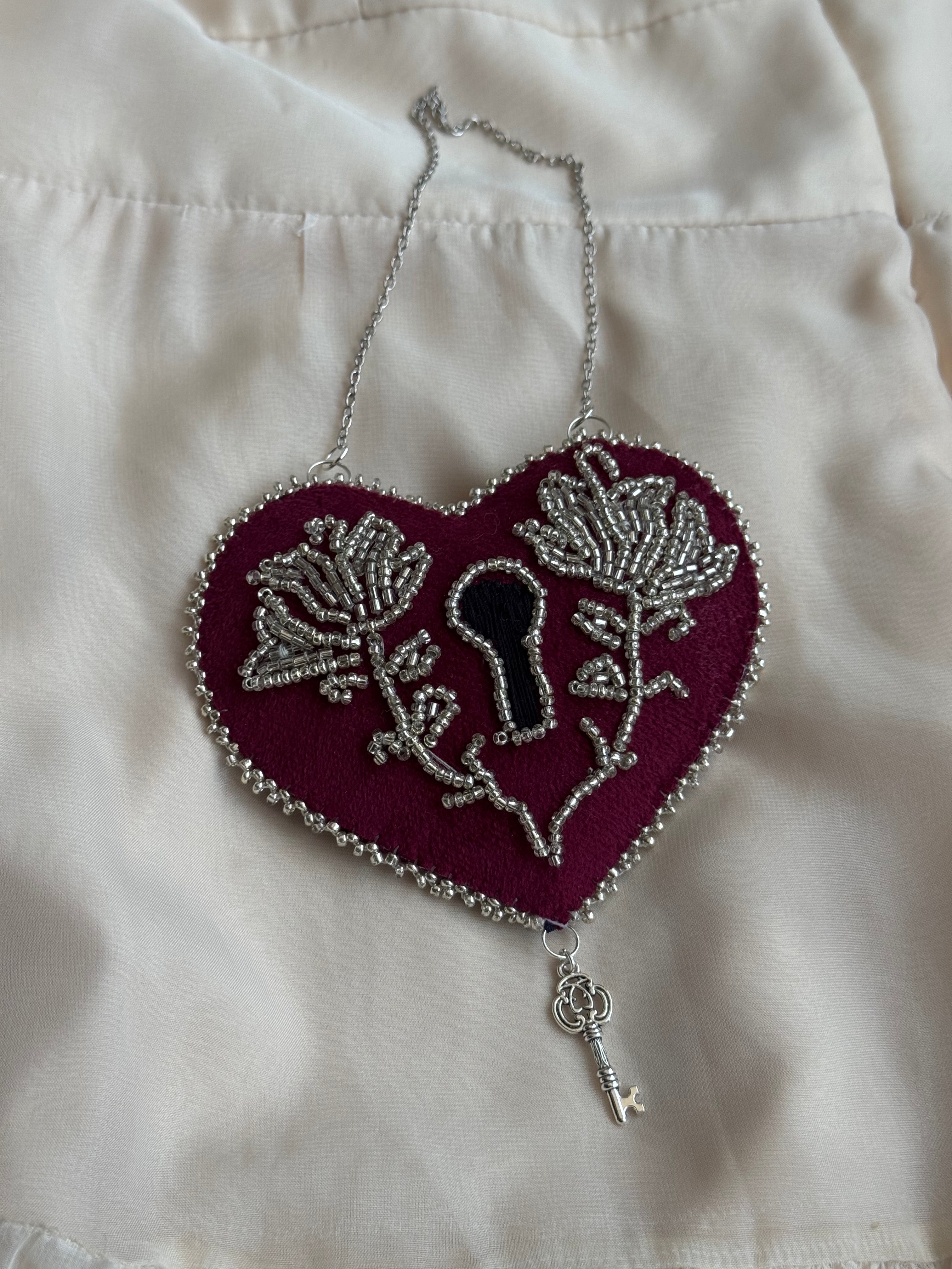Decorative heart shaped, lock-style wall hanging with ornate silver floral design on a white satin background