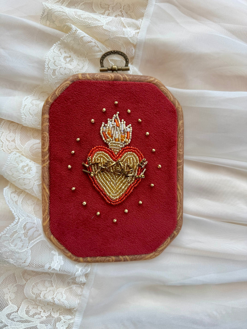Embroidered wall hanging with a sacred heart design on velvet, framed in a wood-look frame with metal hanging, on a white fabric surface.