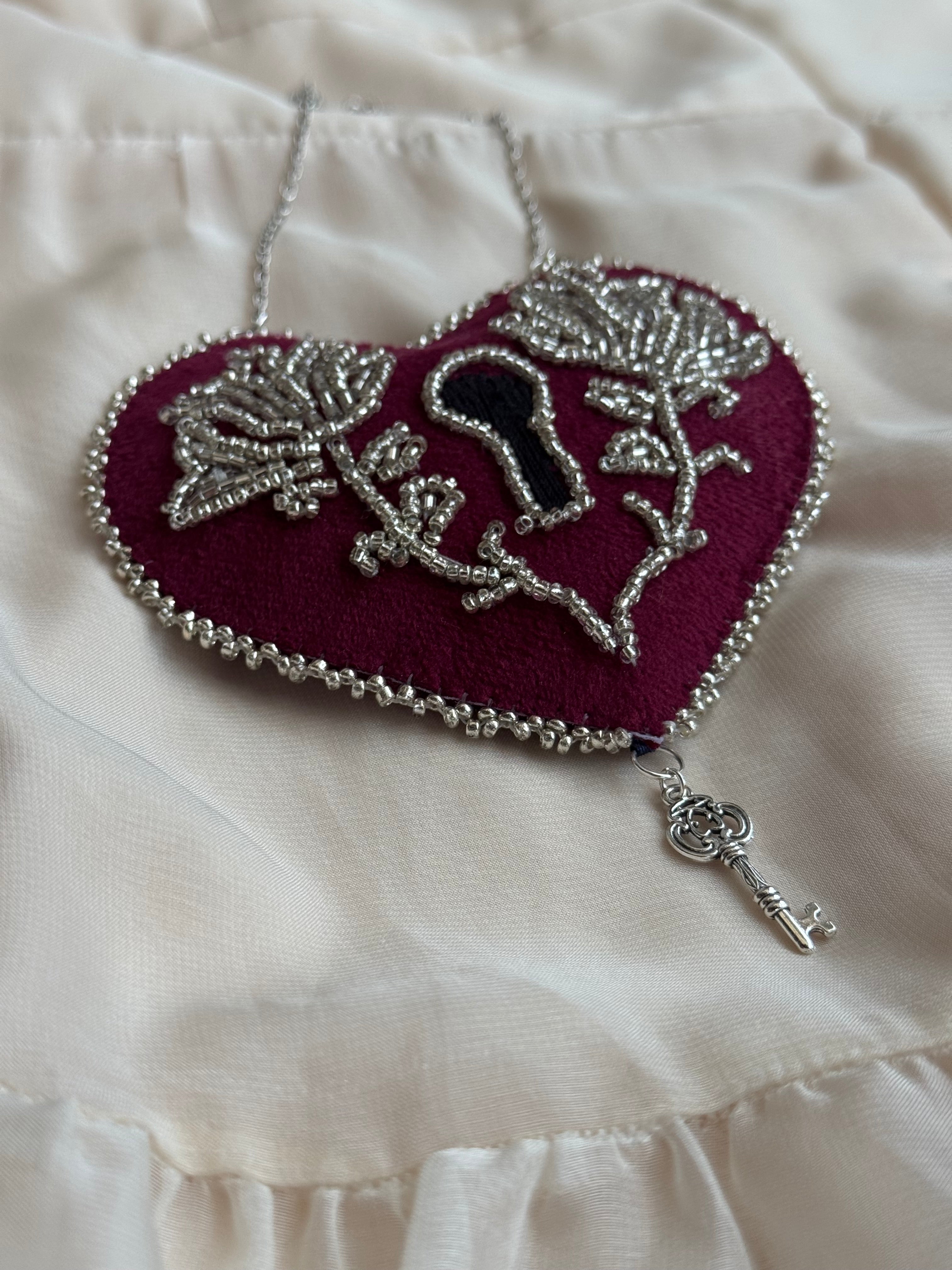 Decorative heart shaped, lock-style wall hanging with ornate silver floral design and dangling silver key charm on a white satin background