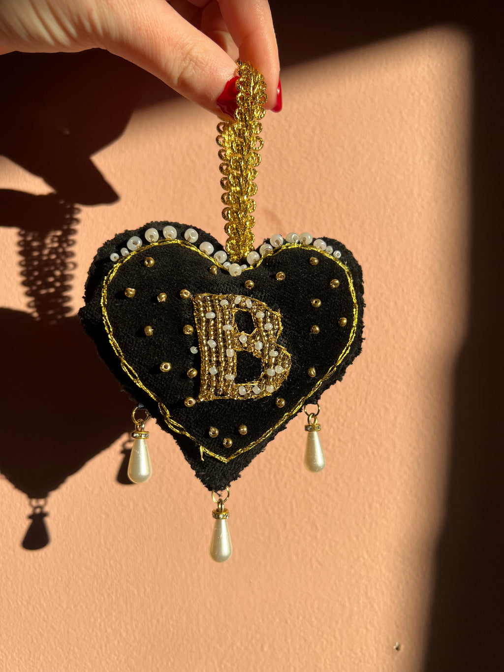 Decorative black heart ornament with gold accents and pearl details, inspired by Anne Boleyn's 'B' necklace, held by a hand on a beige background