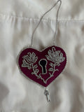 Decorative heart shaped, lock-style wall hanging with ornate silver floral design on a white satin background