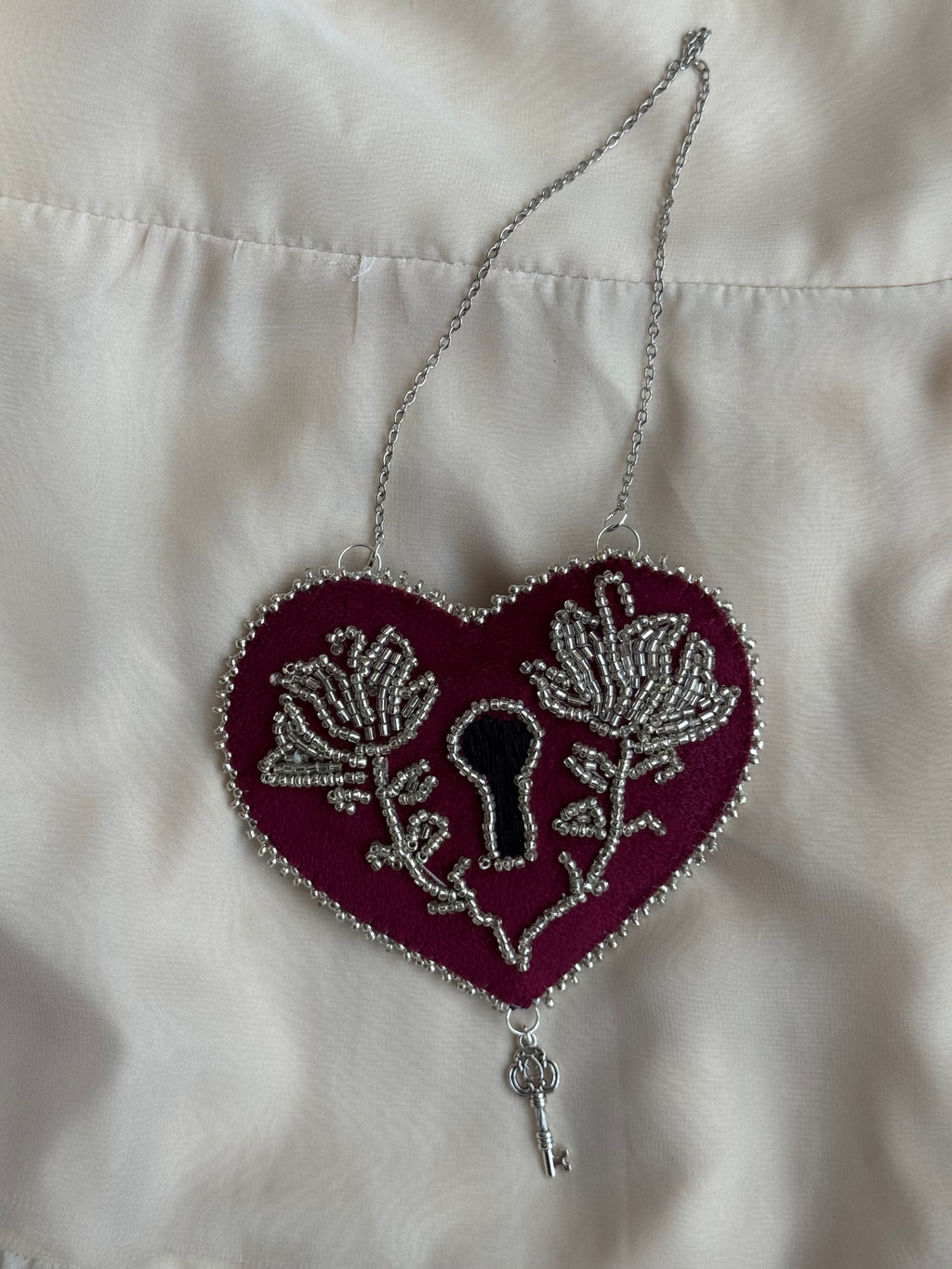 Decorative heart shaped, lock-style wall hanging with ornate silver floral design on a white satin background