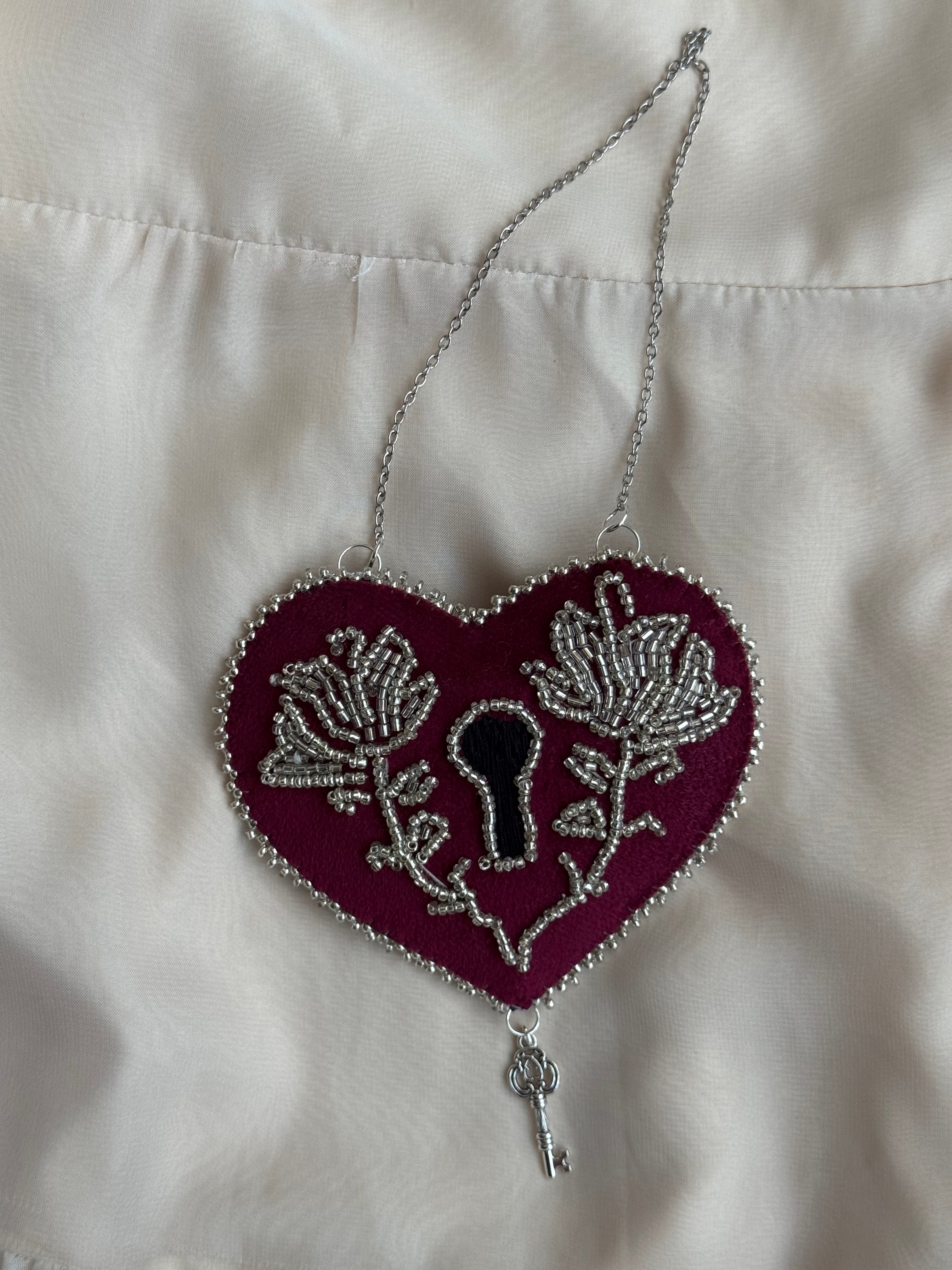 Decorative heart shaped, lock-style wall hanging with ornate silver floral design on a white satin background