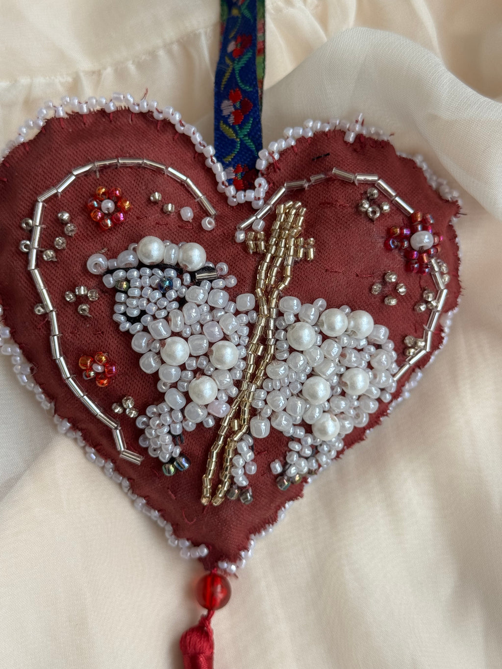 Heart-shaped ornament with beadwork shaping a sacred lamb on a white background with cross, floral hanger and tassle