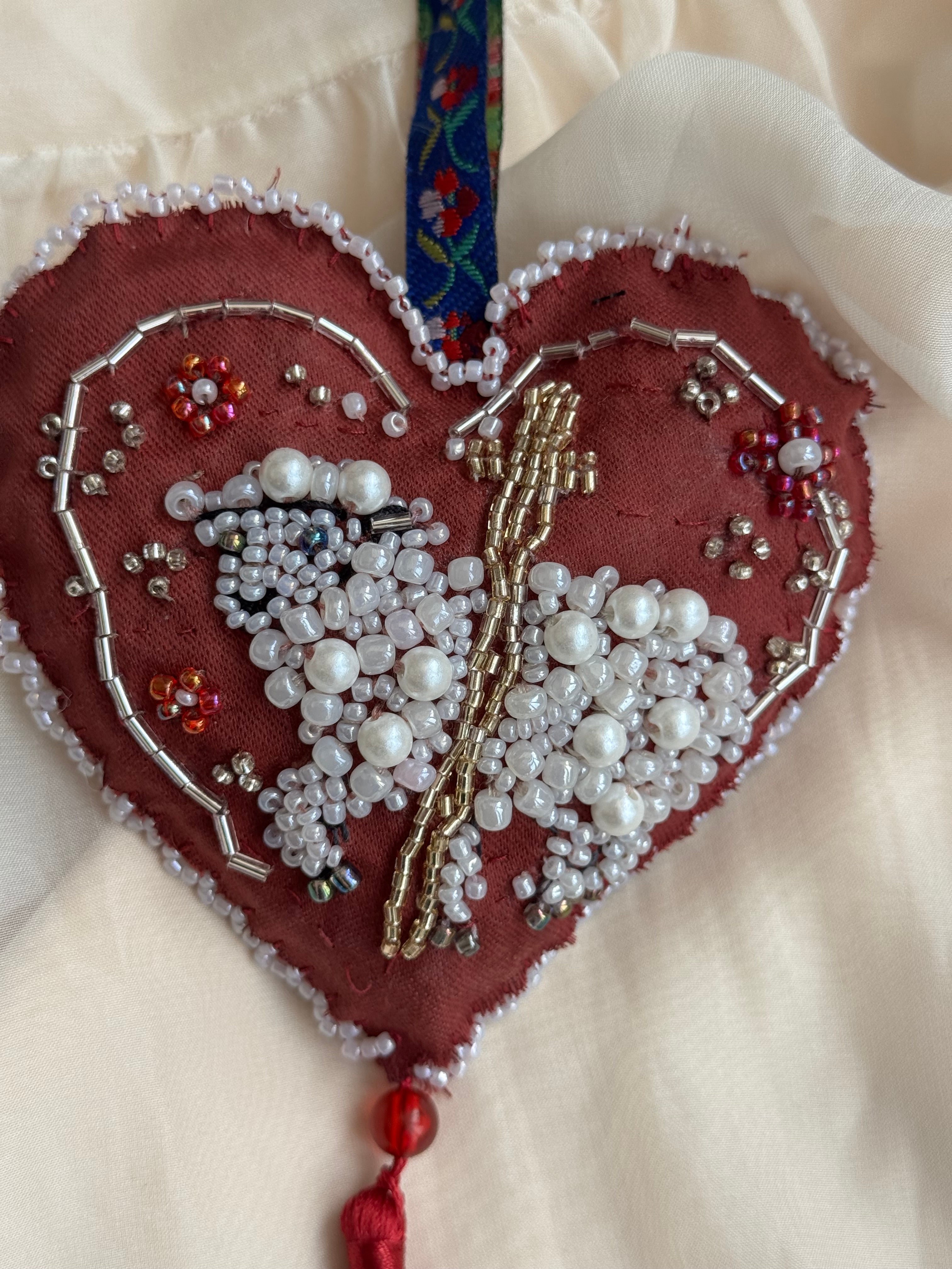 Heart-shaped ornament with beadwork shaping a sacred lamb on a white background with cross, floral hanger and tassle