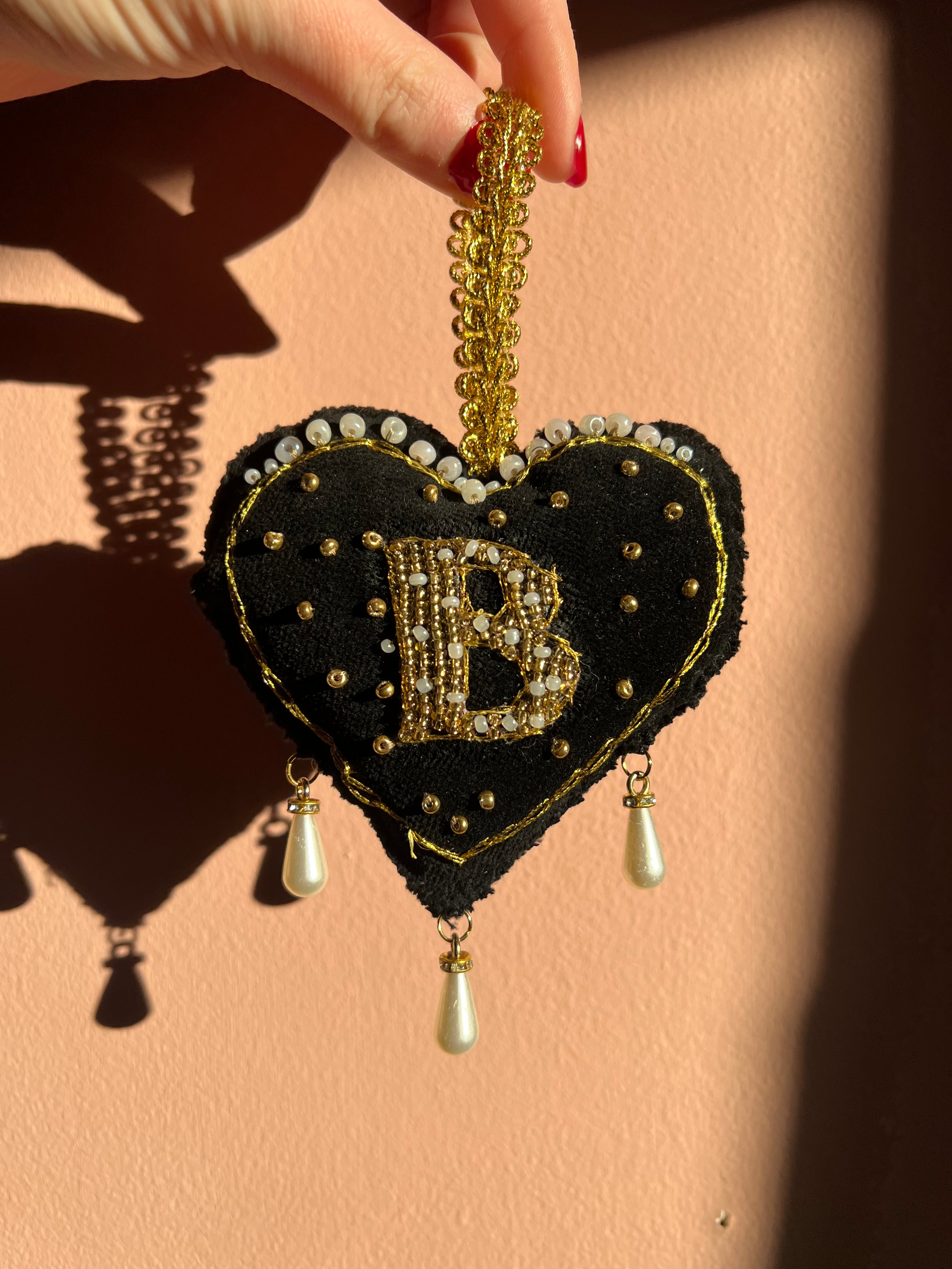 Decorative black heart ornament with gold accents and pearl details, inspired by Anne Boleyn's 'B' necklace, held by a hand on a beige background