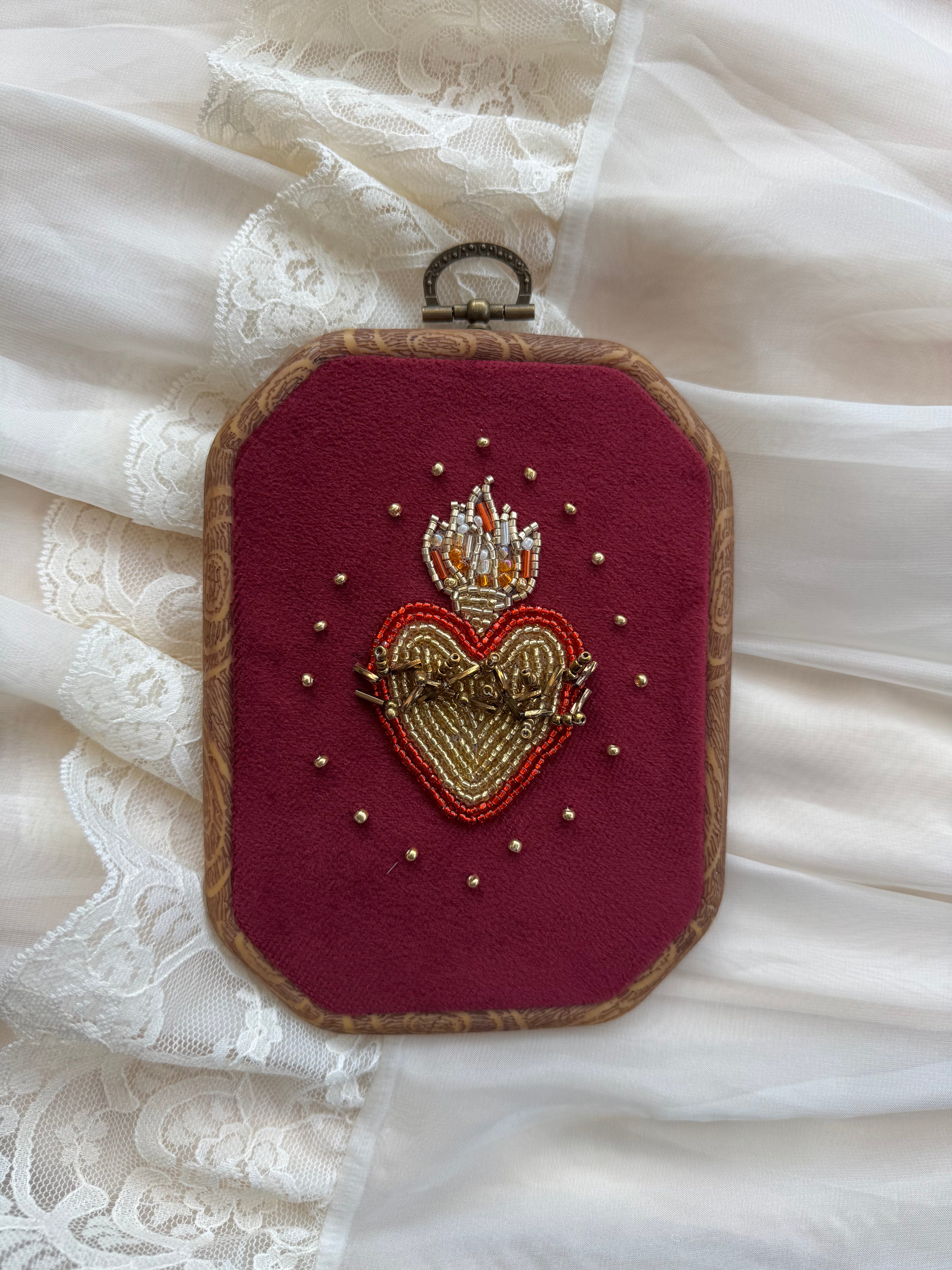 Embroidered wall hanging with a sacred heart design on velvet, framed in a wood-look frame with metal hanging, on a white fabric surface.