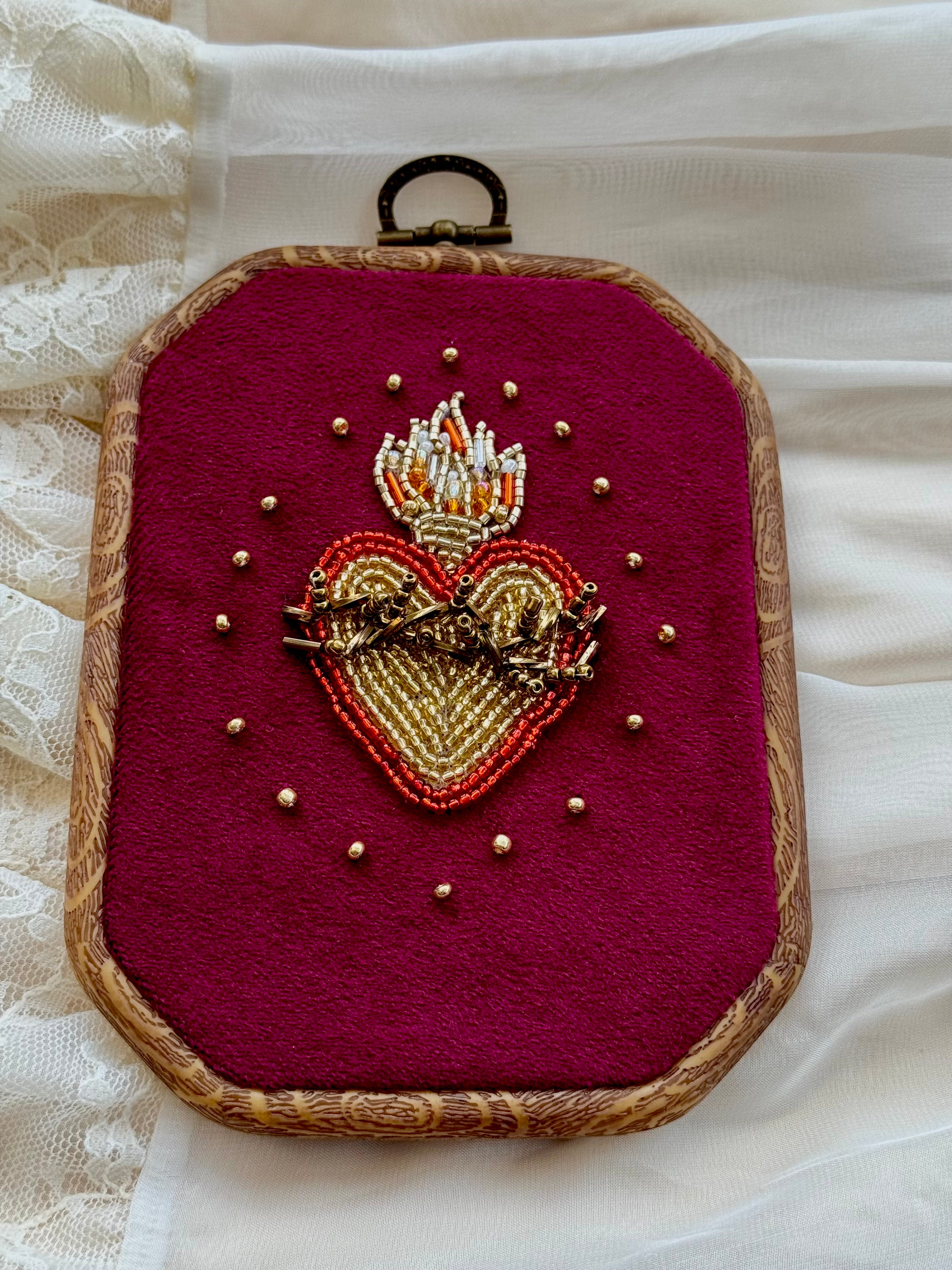 Embroidered wall hanging with a sacred heart design on velvet, framed in a wood-look frame with metal hanging, on a white fabric surface.