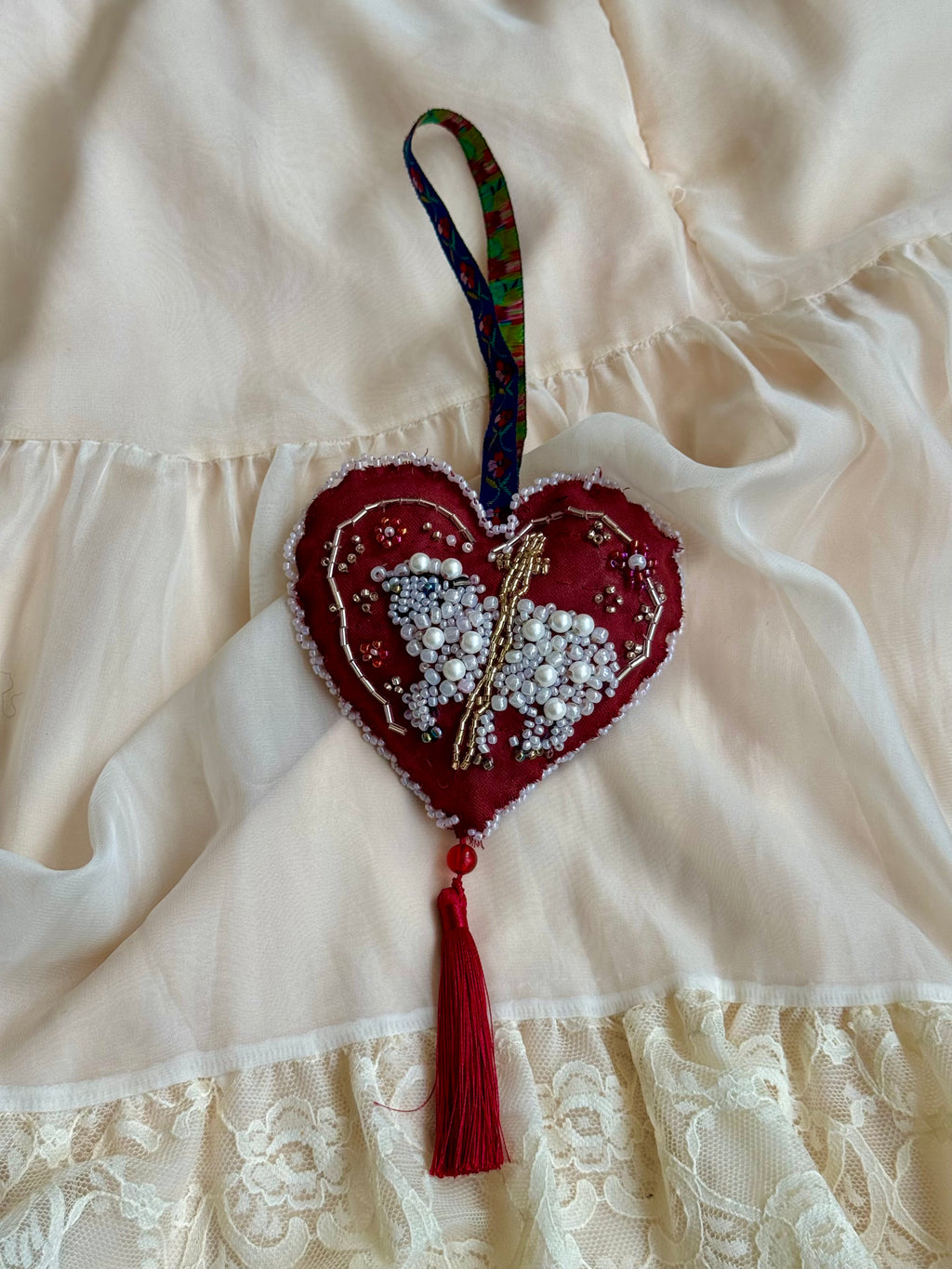 Heart-shaped ornament with beadwork shaping a sacred lamb on a white background with cross, floral hanger and tassle