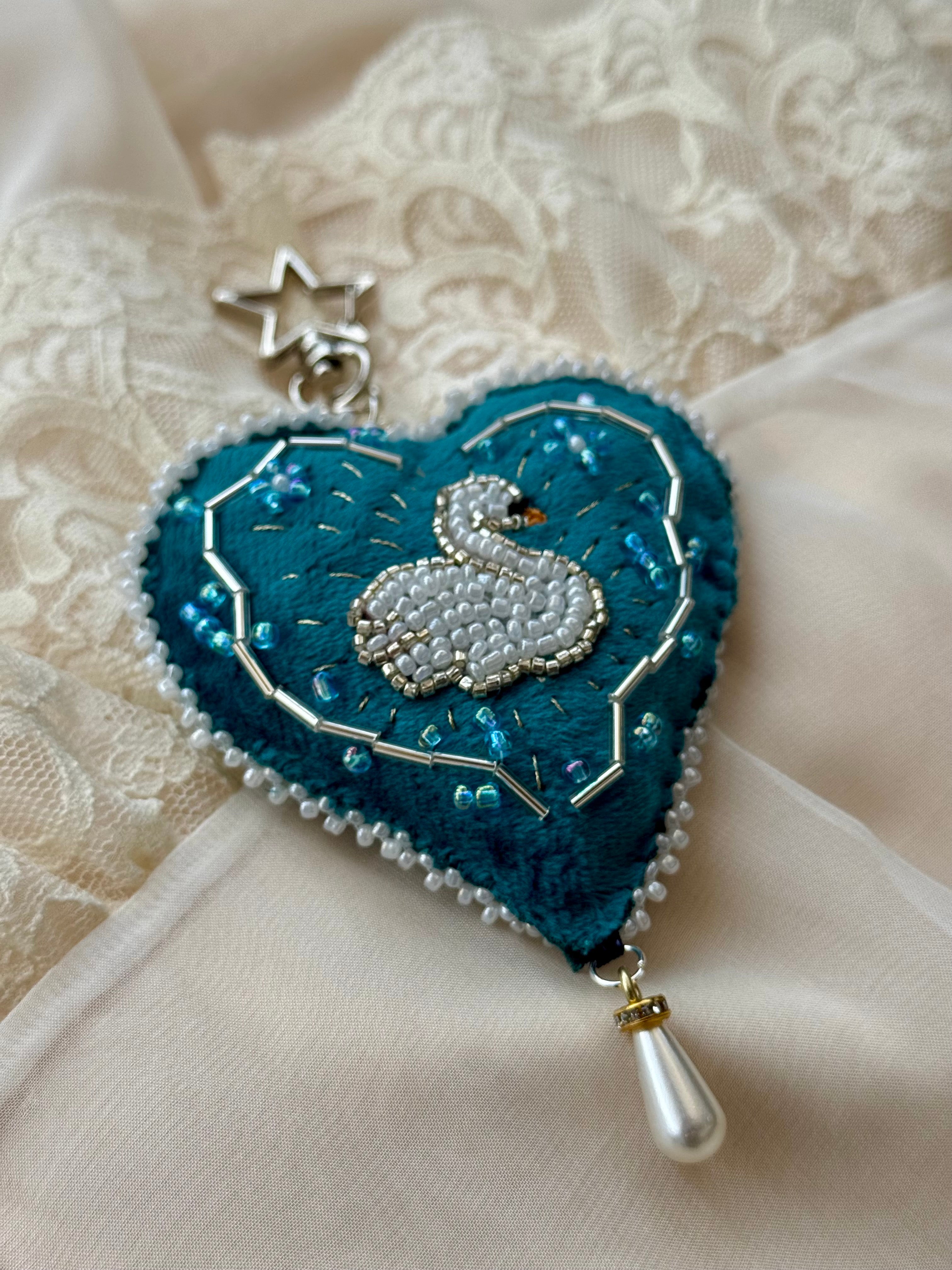 Heart-shaped embroidered pendant with a beaded swan design on a textured fabric background