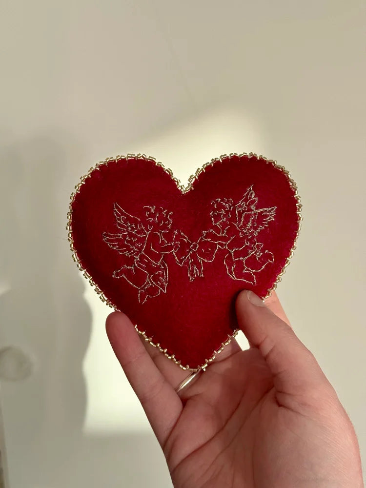 Red heart-shaped cherub embroidered pillow on lace fabric held by a hand against a neutral background