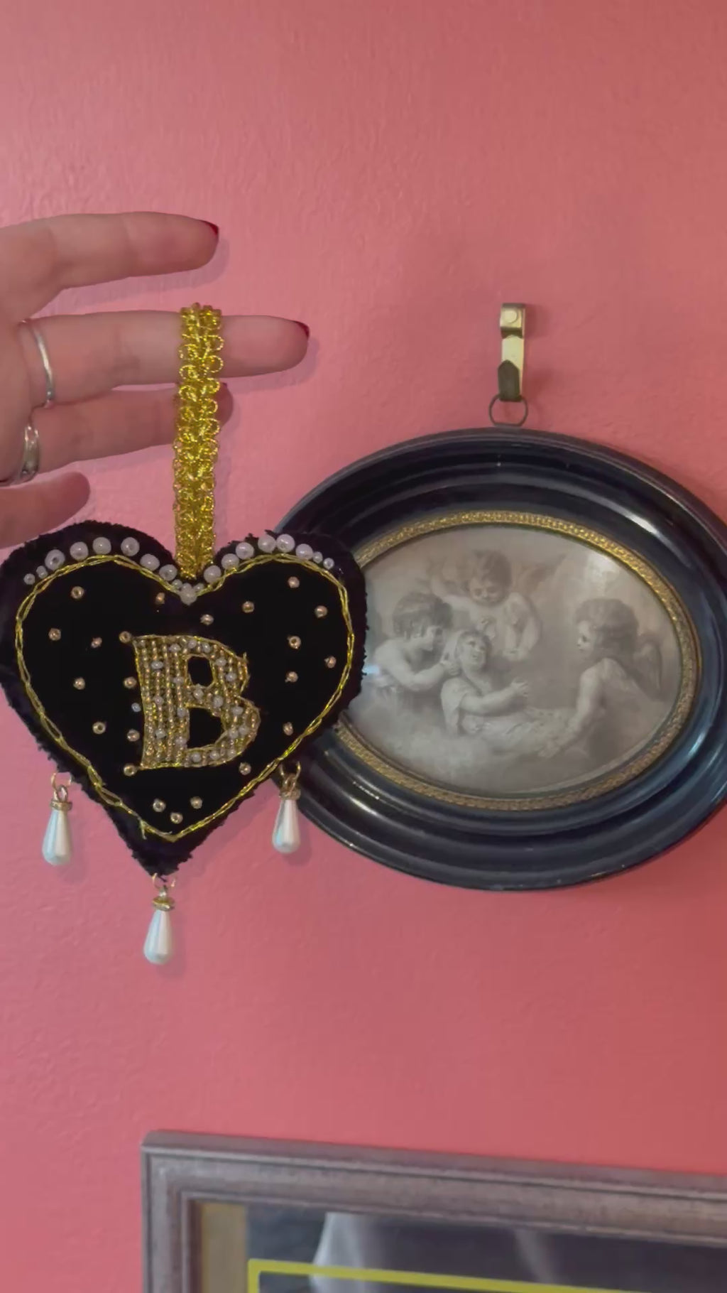 Decorative black heart ornament with gold accents and pearl details, inspired by Anne Boleyn's 'B' necklace, held by a hand on a pink background next to a vintage photo of cherubs.