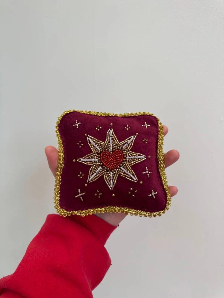 Hand holding a small embroidered pillow with a star and heart design on a plain background