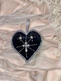 Heart shaped black velvet heart ornament with silver trim and hanger and a cross pendant in the middle on a satin and lace background