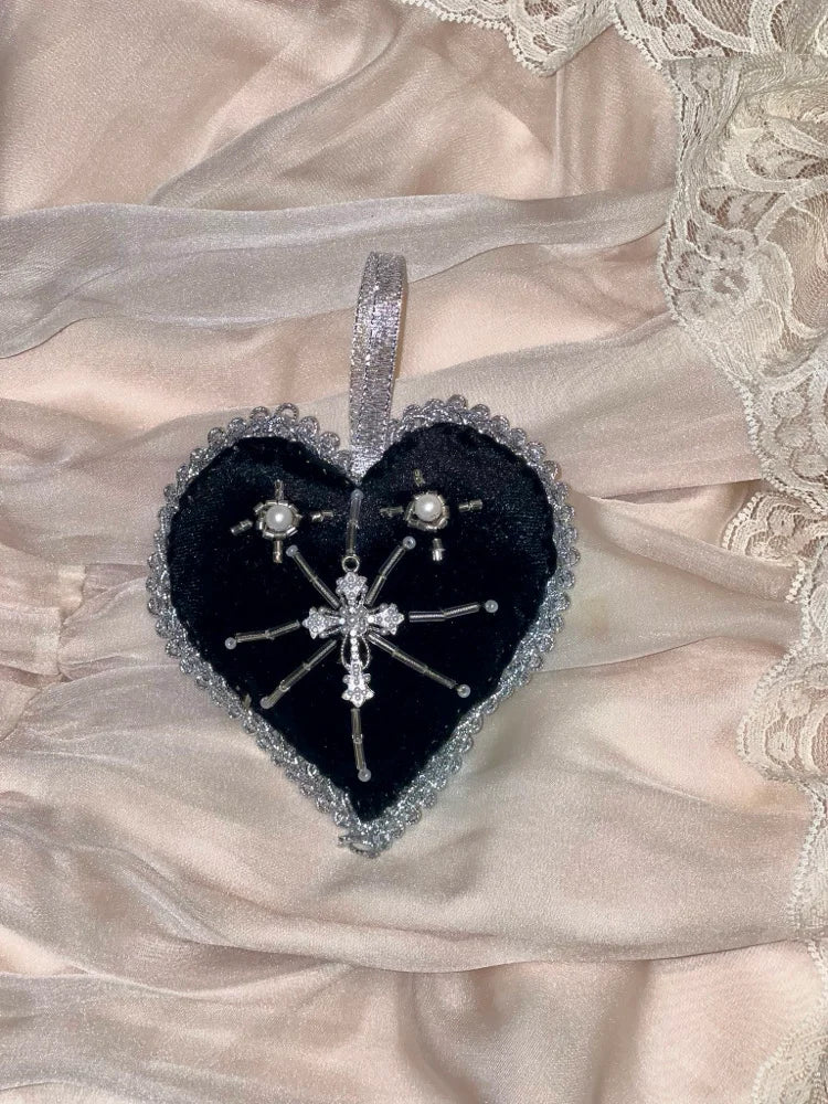 Heart shaped black velvet heart ornament with silver trim and hanger and a cross pendant in the middle on a satin and lace background