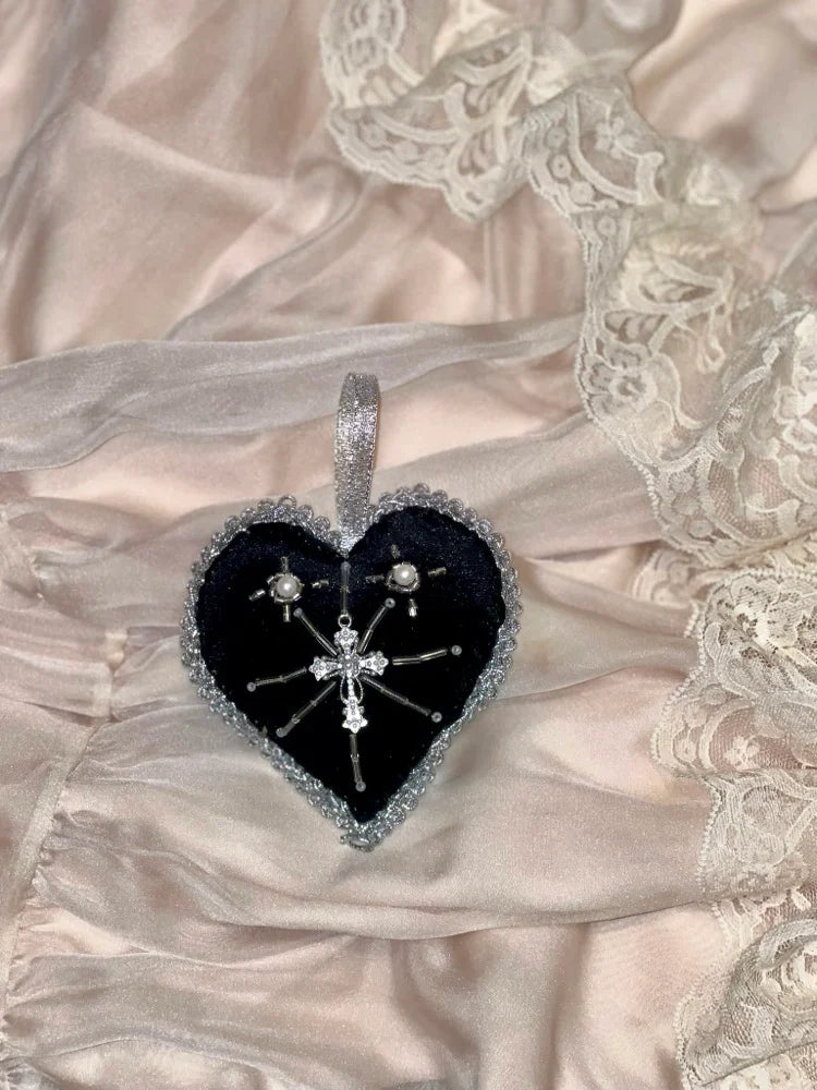 Heart shaped black velvet heart ornament with silver trim and hanger and a cross pendant in the middle on a satin and lace background